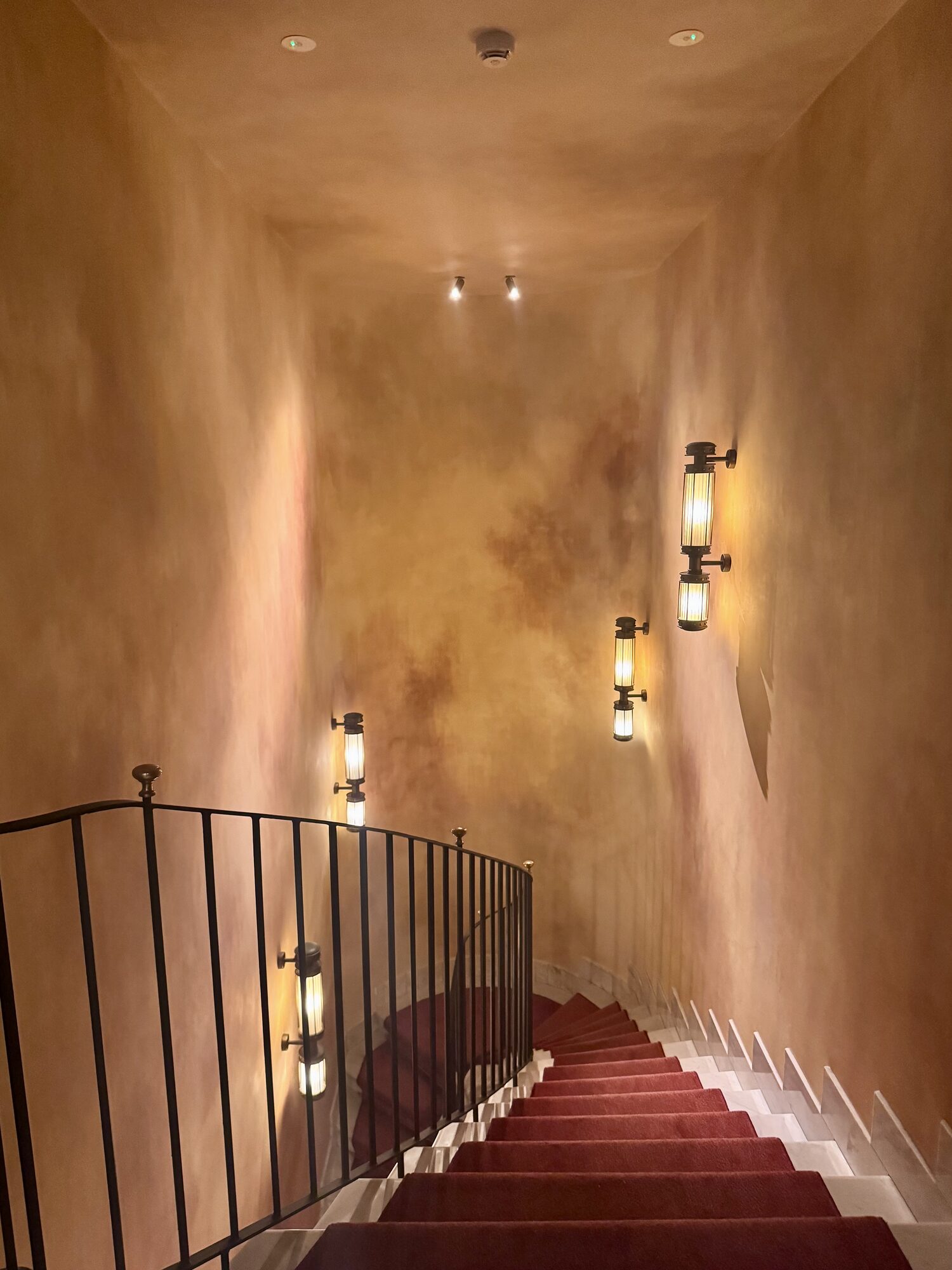 A warm-plaster staircase with iron rail and wall sconces descending over red carpet