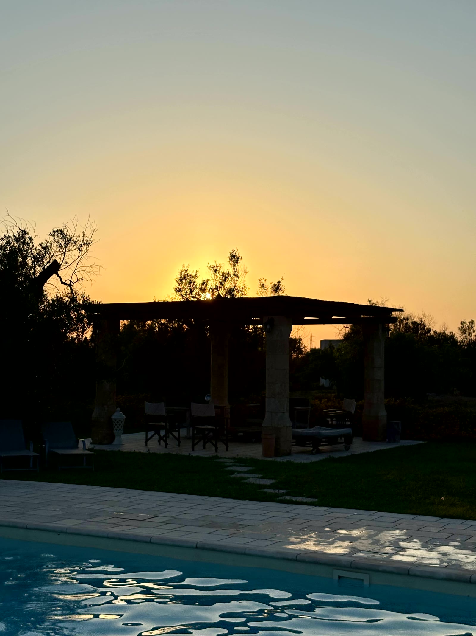 A stone pergola at sunset beyond the pool, the sky going gold behind the olives